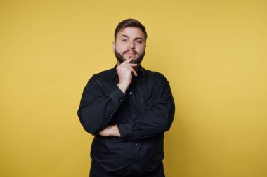 Sakallı bir adam sarı bir duvarın önünde duruyor. Eli çenesinde ve düşünceli biri gibi görünüyor. Işık parlak ve bir stüdyo ayarı..