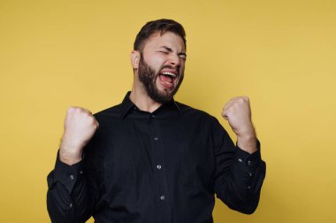 Bir adam sarı bir duvara karşı duruyor. Yumruklarını kaldırır ve heyecanla bağırır. Yüzünde mutluluk ve kutlama ifadesi var. Sahne neşeli bir anı yakalar..