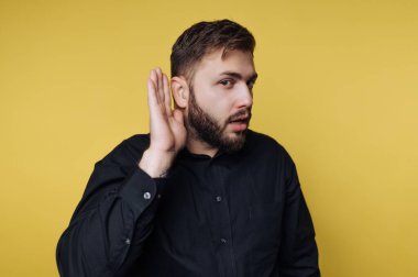 Bir adam elini kulağına dayayıp, dikkatle dinlediğini gösterir. Onun ifadesi ilgi gösteriyor. Arka plan parlak sarı..