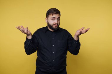 Elleri havada, belirsizlik jesti yapan bir adam. Siyah bir gömlek giyiyor ve açık renk bir sakalı var. Arka plan parlak sarı. Sahne kafa karışıklığını yakalar..