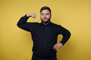 Stüdyoda parlak sarı bir geçmişi olan bir adam var. Diğer elini kalçasında tutarken kol kaslarını esnetiyor. Yüzünde hafif bir gülümseme var ve siyah bir gömlek giyiyor..