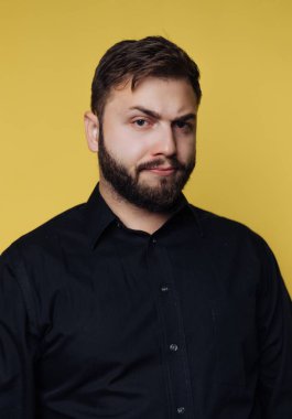 Bir adam parlak sarı bir arka plana karşı duruyor. Siyah bir gömlek giyiyor ve yüzünde ciddi bir ifade var. Sakalı onun belirgin görünümüne ekleniyor. Doğrudan izleyiciye bakıyor..