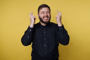 Bir adam parlak bir gülümsemeyle sarı bir duvarın önünde duruyor. Parmaklarını çapraz yapar ve umut verici bir ifade gösterir. Ayar parlak ve neşeli görünüyor..