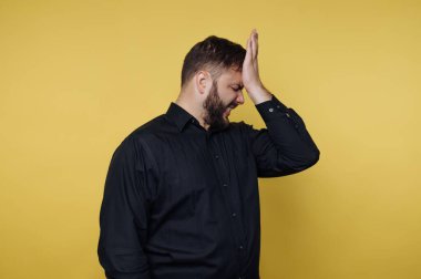 Bir adam sarı bir zemine karşı duruyor. Alnına tek eliyle dokunduğunda hayal kırıklığına uğramış gibi görünüyor. Yüzü, stresli bir durumla karşı karşıya olduğunu gösteriyor..