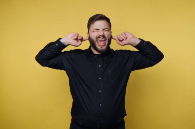 Kollarını yan taraflarına koyup kulaklarını kapatan bir adam. O, parlak sarı bir arkaplan önünde iken yüzünde neşeli bir ifadeye sahiptir. Sahne bir eğlence ve mizah anını yakalar..