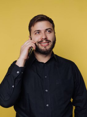 Telefonla konuşan bir adam var. Sakalı ve gülüşü var. Gömleği siyah. Arka plan sarı. Bu konuşma sırasında mutlu görünüyor..