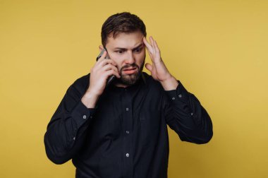 Bir adam parlak sarı bir arka plana karşı duruyor. O, bir eliyle kulağına bir telefon tutarken diğer eli başının üstündedir. Konuşurken endişeli ve şaşırmış görünüyor..