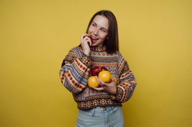 Sarı bir duvarın önünde elinde meyvelerle duran bir kadın. Gülümsüyor ve meyvelerden birini ısırıyor. Kapalı alanda geçen neşeli ve neşeli bir an..