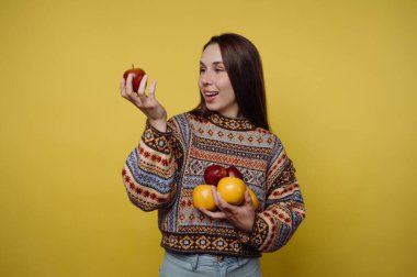 Sarı arka planda bir kadın duruyor. Bir elinde bir elma, diğer elinde birkaç elma tutuyor. Mutlu görünüyor ve bu andan zevk alıyor..