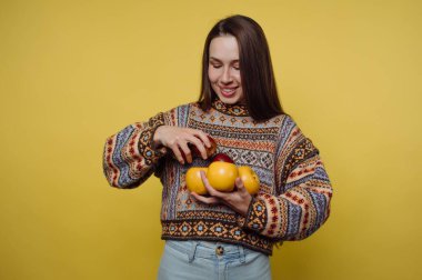 Elmalar ve portakallar da dahil birçok meyve tutan bir kadın gülümseyerek ayakta duruyor. Kalıplı bir süveter giyiyor ve sarı bir arkaplanda poz veriyor..