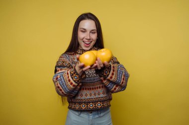 Genç bir kadın parlak sarı bir duvarın önünde duruyor. Elinde üç portakal tutarken gülümsüyor. Dekor basit ve renkli, canlı bir atmosfer yaratıyor..