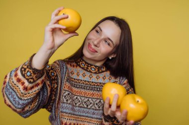 Genç bir kadın sarı arka planda duruyor. Üç portakal tutuyor ve birine yakından bakıyor. Elindeki meyvelerle meşgul olduğunda mutlu görünüyor..