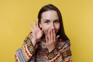 Uzun saçlı bir kadın eliyle ağzını kapatır ve gülümser. Kalıplı bir süveter giyiyor ve koyu sarı bir arka planın önünde duruyor..