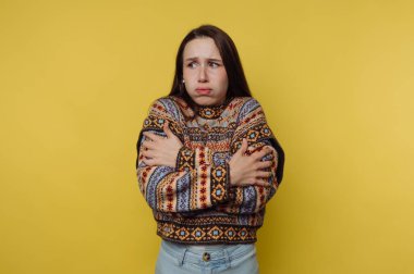 Sarı arka planda bir kadın var. Kollarını kavuşturmuş ve belirsiz bir ifade sergiliyor. Kazağın renkli bir deseni var..