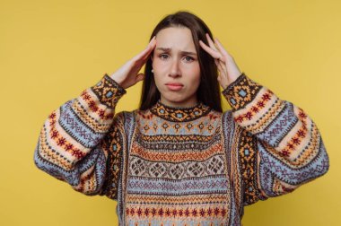 Elleri başının üstünde duran genç bir kadın, ezilme belirtileri gösteriyor. Renkli bir süveter ve endişeli bir ifade takınıyor. Arka plan parlak sarı..