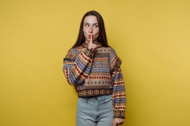 Sarı arka planda bir kadın duruyor. O, desenli bir kazak giyer ve parmağını dudaklarına koyar. Sessizlik ya da gizlilik istiyor gibi görünüyor. Neşeli bir şekilde..