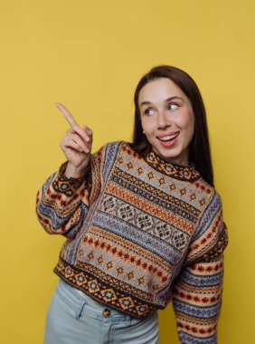 Genç bir kadın parlak sarı bir arka planın önünde duruyor. Kocaman gülümsüyor ve tek parmakla işaret ediyor. Renkli bir süveter ve açık renk pantolon giyiyor..
