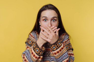 Bir kadın elleriyle ağzını kapatarak şaşkınlığını ifade eder. Uzun saçları var ve renkli bir kazak giyiyor. Arkaplan parlak sarı, gündüz neşeli bir hava yaratıyor..