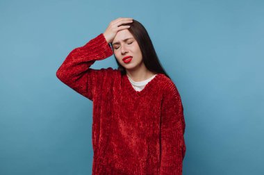 Bir kadın mavi bir duvarın karşısında duruyor, bir eli alnında, yüzü asık. Kırmızı bir kazak giyiyor ve rahatsızlığı ya da ağrısı var gibi görünüyor, muhtemelen stresli..