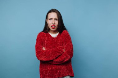 Kollarını kavuşturmuş şaşırmış ve kafası karışmış bir kadın. Uzun saçları var ve kırmızı bir kazak giyiyor. Mavi arkaplan onun ifadesine karşıtlık ekler.