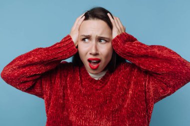 Bir kadın iki eliyle kulaklarını kapatırken stres belirtileri gösterir. O, kırmızı bir kazak giyerken endişeli görünüyor. Ayarlar mavi arkaplan ile basit.