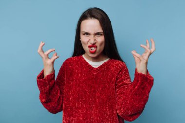 Bir kadın kırmızı bir kazak giyerken güçlü duygular gösterir. Elleri havada, sert bir yüz ifadesi takınıyor. Basit mavi arkaplan güçlü duruşuna odaklanmasını sağlıyor..