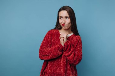 Genç bir kadın elleri ve kaşları çatık bir şekilde ayakta duruyor. O, kararsız görünüyor ve kırmızı bir kazak giyiyor. Arkaplan sade mavi renkte..