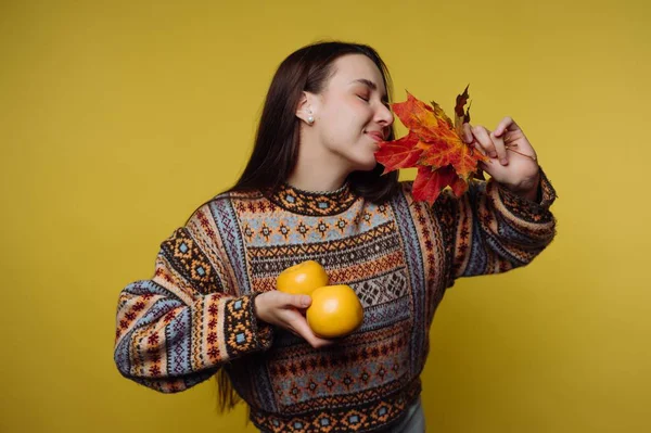 Bir elinde iki portakal, diğer elinde renkli sonbahar yaprakları tutan bir kadın. Anın tadını çıkarırken gülümsüyor. Arka plan parlak sarı, sahneyi vurguluyor..