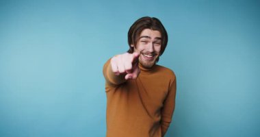 Bir kişi mavi bir arka planın önünde duruyor, gülümsüyor ve işaret ediyor. Etkileşim halindeyken neşe ve dostluk gösteriyor. Her kare bir heyecan anı yakalıyor..