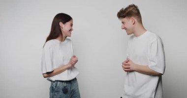 İki genç yüz yüze durup sohbet ediyorlar. Konuşurken gülümser ve hareket ederler. Aydınlık ve basit bir ortam. Onların etkileşimlerine ve ifadelerine odaklan..