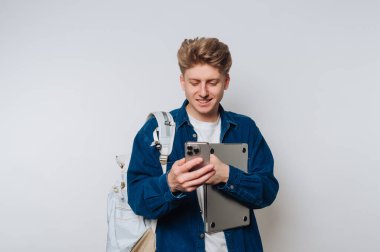 Genç bir adam neşeli bir gülümsemeyle ayakta duruyor, bir elinde akıllı bir telefon, diğer elinde bir dizüstü bilgisayar. Mavi bir ceket giyiyor, gündelik tarzına modern bir dokunuş katıyor..