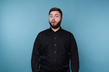 Bir adam canlı, mavi bir stüdyoda duruyor, ağzı şokta. Siyah gömlekli, şaşırmış ifadesi izleyicileri cezbediyor, merak ve entrika uyandırıyor..