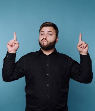 Siyah gömlekli şık bir adam çarpıcı mavi bir duvara karşı duruyor. Kendinden emin bir şekilde parmaklarını yukarı kaldırıyor. Çekicilik ve neşe saçıyor. Seyirciyi kendine çekiyor..