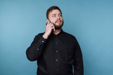 Sakallı bir adam açık mavi bir duvara karşı duruyor, telefonunu kulağına dayıyor. Bir sohbete dalmış gibi görünüyor, özgüven ve odaklanma yayıyor..