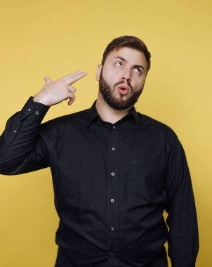 Sakallı genç bir adam kendini parmaklarıyla vuruyormuş gibi yaparak eğlenceli bir poz veriyor. Siyah bir gömlek giyiyor ve parlak sarı bir arka plana karşı duruyor, neşeli bir enerji gösteriyor..