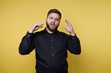 Siyah tişört giyen sakallı bir adam, parlak sarı bir duvarın önünde düşüncelerini ifade ederken tutkulu bir şekilde el hareketi yapar ve çekici bir atmosfer yaratır..