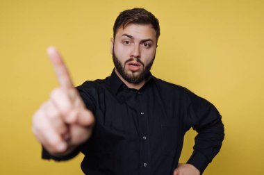 Bir adam, bir eli havada, özgüvenle ayakta duruyor ve güçlü bir noktayı ifade ediyor. Parlak sarı bir arka plana karşı siyah bir gömlek giyen o, iddialı duruşuyla dikkati çeker..