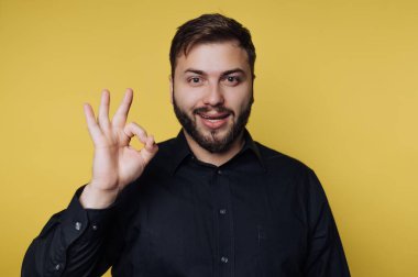 Neşeli bir gülümseme ile, siyah gömlekli genç bir adam iyi bir işaret gösterir. Canlı sarı arkaplan, bu canlı ana sıcaklık katıyor. Bu da onu davetkar ve dost canlısı yapıyor..