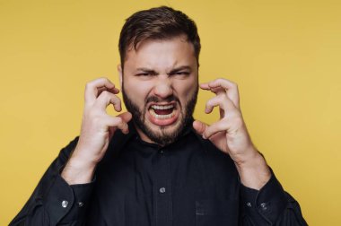 Kalın sakallı bir adam, parmaklarını sıkıca sıkarken bakışlarını öne doğru sabitleyen yoğun bir hayal kırıklığı gösterir. İfadesi öfke saçıyor, canlı sarı bir arkaplanda yakalanmış..