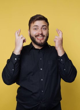 Siyah gömlek giyen arkadaş canlısı bir adam şans dilemek için şans dilerken geniş bir gülümseme sergiliyor. Parlak sarı arka plan onun umut verici jestine enerji katıyor. Bu an neşeyi ve beklentiyi yakalar..