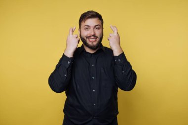 Neşeli bir adam, umut ve şans dilemek için parmaklarını şakayla çaprazlayarak, canlı sarı bir zemin üzerinde duruyor. Gülüşü olumlu, anı neşeli kılıyor..