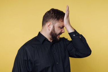 Sakallı bir adam, alnını tutarken hayal kırıklığı belirtileri gösteren parlak sarı bir duvarın önünde duruyor. Siyah bir gömlek giyiyor, hayal kırıklığıyla ilgili bir anı yakalıyor..