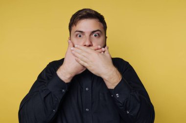 Şok olmuş genç bir adam, elleri şokla ağzını kapatıyor. Geniş gözleri ve parlak siyah gömleği parlak sarı arkaplanla keskin bir tezat oluşturarak yoğun bir görsel etki yaratıyor..