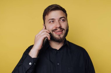 Neşeli genç adam telefonda konuşuyor, parlak bir gülümseme sergiliyor. Günlük siyah gömleği, hayat dolu sarı arkaplanla çok güzel zıtlaşıyor ve neşeli bir atmosfer yaratıyor..