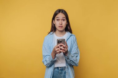 Uzun saçlı genç bir kız canlı sarı bir duvarın önünde duruyor. İnternette biriyle konuşurken akıllı telefonunu şaşırmış bir ifadeyle tutuyor..