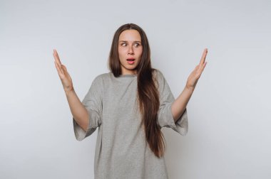 Kolları açık, gözleri açık genç bir kadın şaşırmış bir şekilde ayakta duruyor. Sade, açık renkli bir yerde rahat gri bir gömlek giyerken uzun saçları omuzlarının üzerine düşüyor..