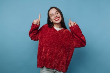 Genç bir kadın sevinçle poz veriyor, sıcak kırmızı bir kazak giyerken büyük bir gülümseme sergiliyor. İki elini de neşeyle kaldırarak eğlenceli ve enerjik bir atmosfer yaratıyor..