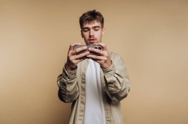 Genç bir adam akıllı telefonunu iki elinde tutarak güvenle ayakta duruyor. Bakışları ekrana sabitlenir ve kendini yumuşak bej bir arka planla çevrili heyecan verici bir mobil oyuna kaptırır..