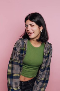 Siyah saçlı bir kız kameraya göz kırparak neşeli tarafını gösteriyor. Yeşil bir bluz ve ekose bir gömlek giyer, büyüleyici pembe bir arkaplan karşısında güvenle durur..