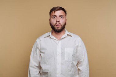 Tarafsız renkli bir odada duran bir adam, gözleri geniş ve ağzı tamamen şokta. Beyaz gömleği, arkasındaki yumuşak, ılık tonlu duvarla çelişiyor. Gerçek bir sürpriz anı yakalıyor..